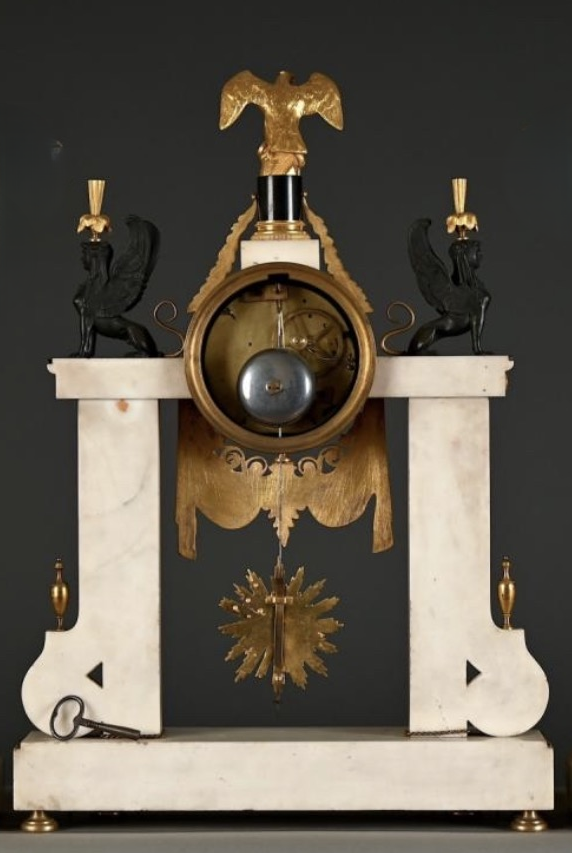 Large Empire-style portico clock (64.5 cm), 19thC. Large clock featuring two marble columns adorned with two caryatids, rosettes, drapery, decorative motifs, two patinated and gilded sphinxes, and an eagle at the top. Sun dial and key included