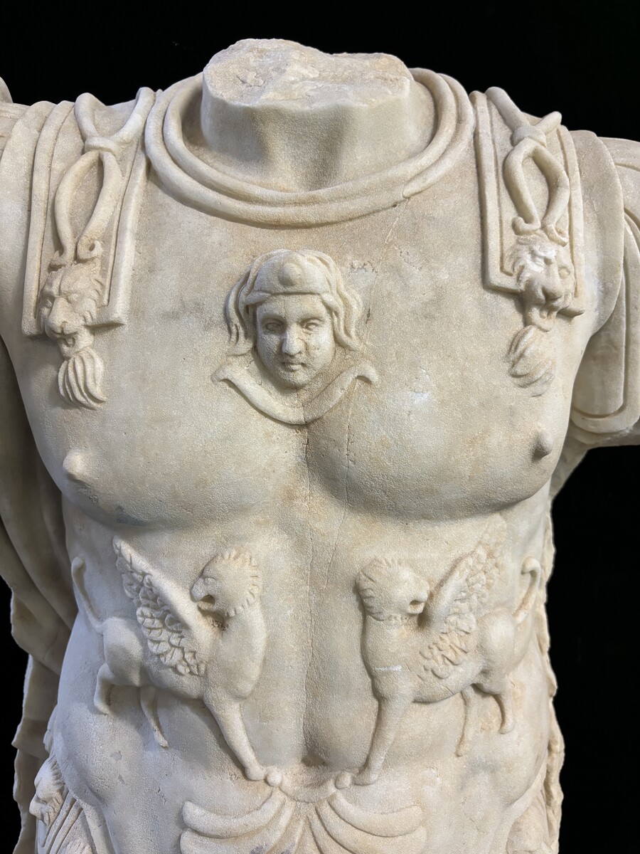 An impressive sculpture of a Roman torso in white marble, exuding the power and beauty of classical antiquity. The detailed breastplate is richly decorated with hand-carved mythological motifs. 