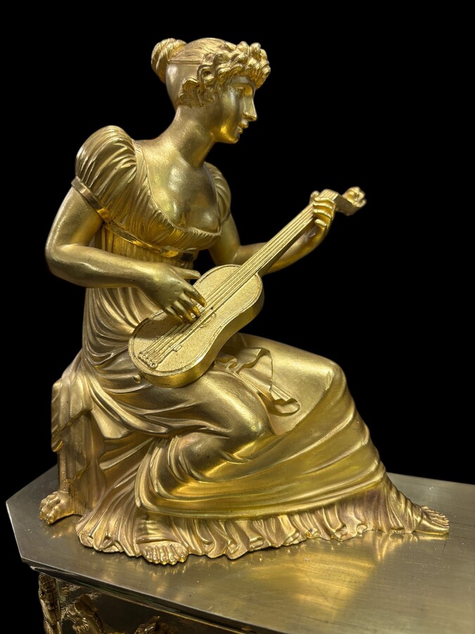 Beautiful Empire period clock in matt and bright gilt bronze depicting a lady playing guitar with an angel. In very good condition, the movement has not been checked. The balance wheel is present. Dimensions: Width: 31.5 cm Height: 43 cm Depth : 13 cm