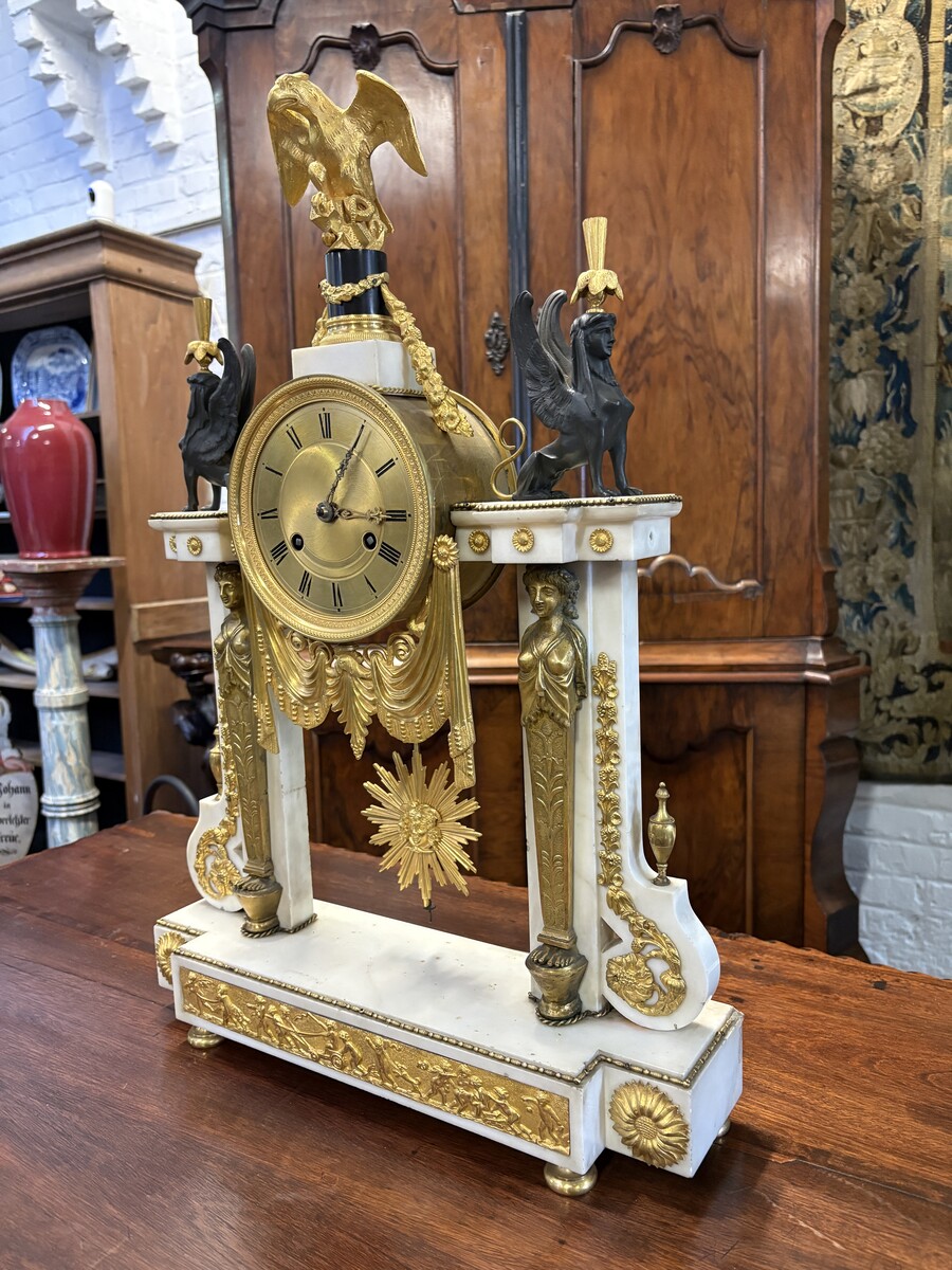 Large Empire-style portico clock (64.5 cm), 19thC. Large clock featuring two marble columns adorned with two caryatids, rosettes, drapery, decorative motifs, two patinated and gilded sphinxes, and an eagle at the top. Sun dial and key included; move