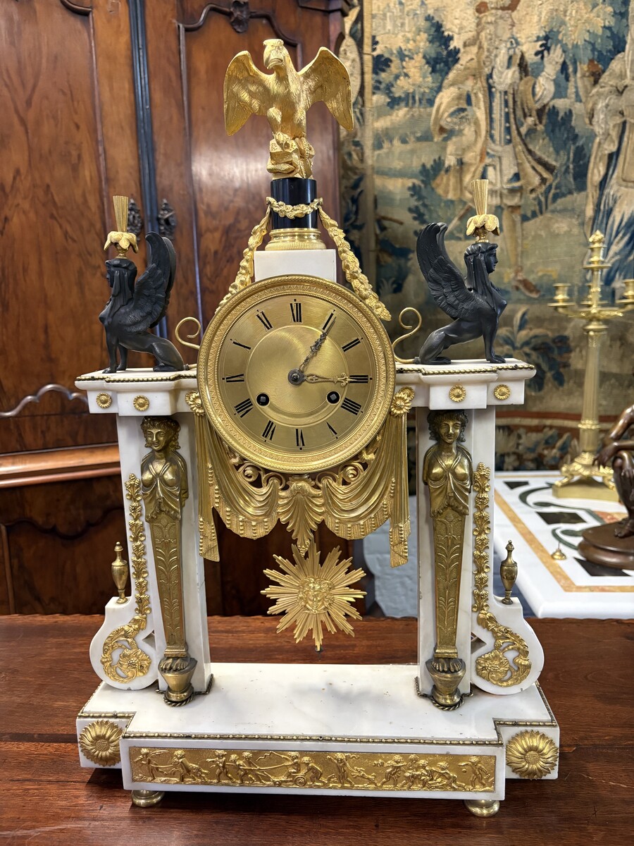 Large Empire-style portico clock (64.5 cm), 19thC. Large clock featuring two marble columns adorned with two caryatids, rosettes, drapery, decorative motifs, two patinated and gilded sphinxes, and an eagle at the top. Sun dial and key included; move