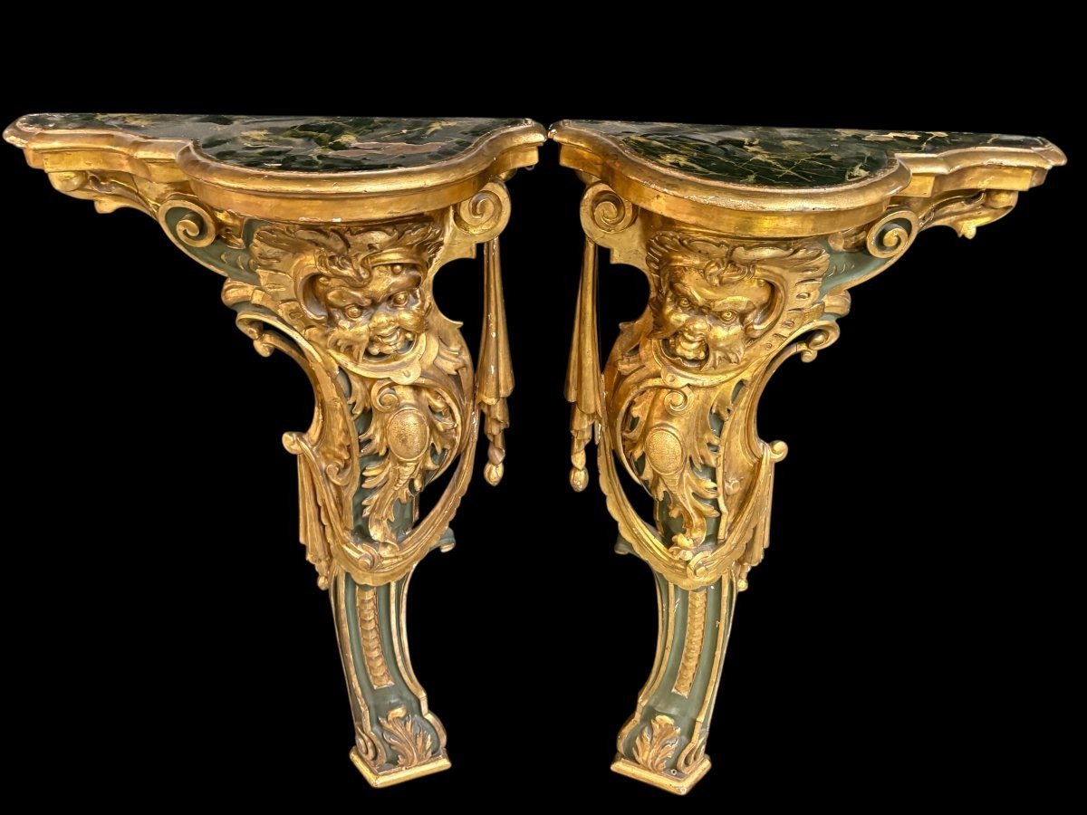 Pair of large, elegant 18th-century Italian wall consoles. Highly decorative consoles in gilded and patinated wood, adorned with a mythological head and decorated with drapery, volutes and ornaments. The top shelf is painted in faux marble.