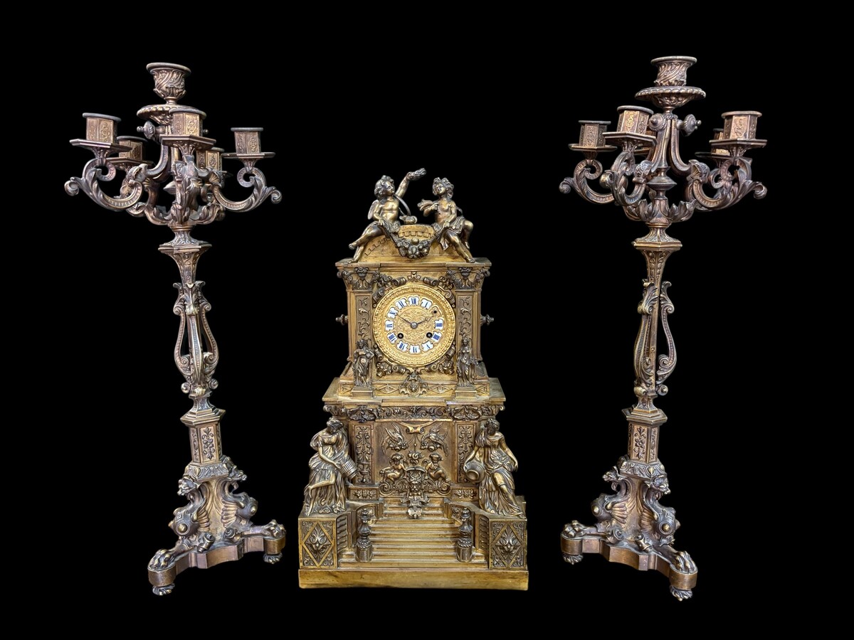 Romantic Napoleon III-style bronze mantelpiece set A mantelpiece set comprising a clock adorned with angels, figures, decorative motifs and a building with a staircase, as well as a pair of candlesticks richly decorated with mythological creatures. 